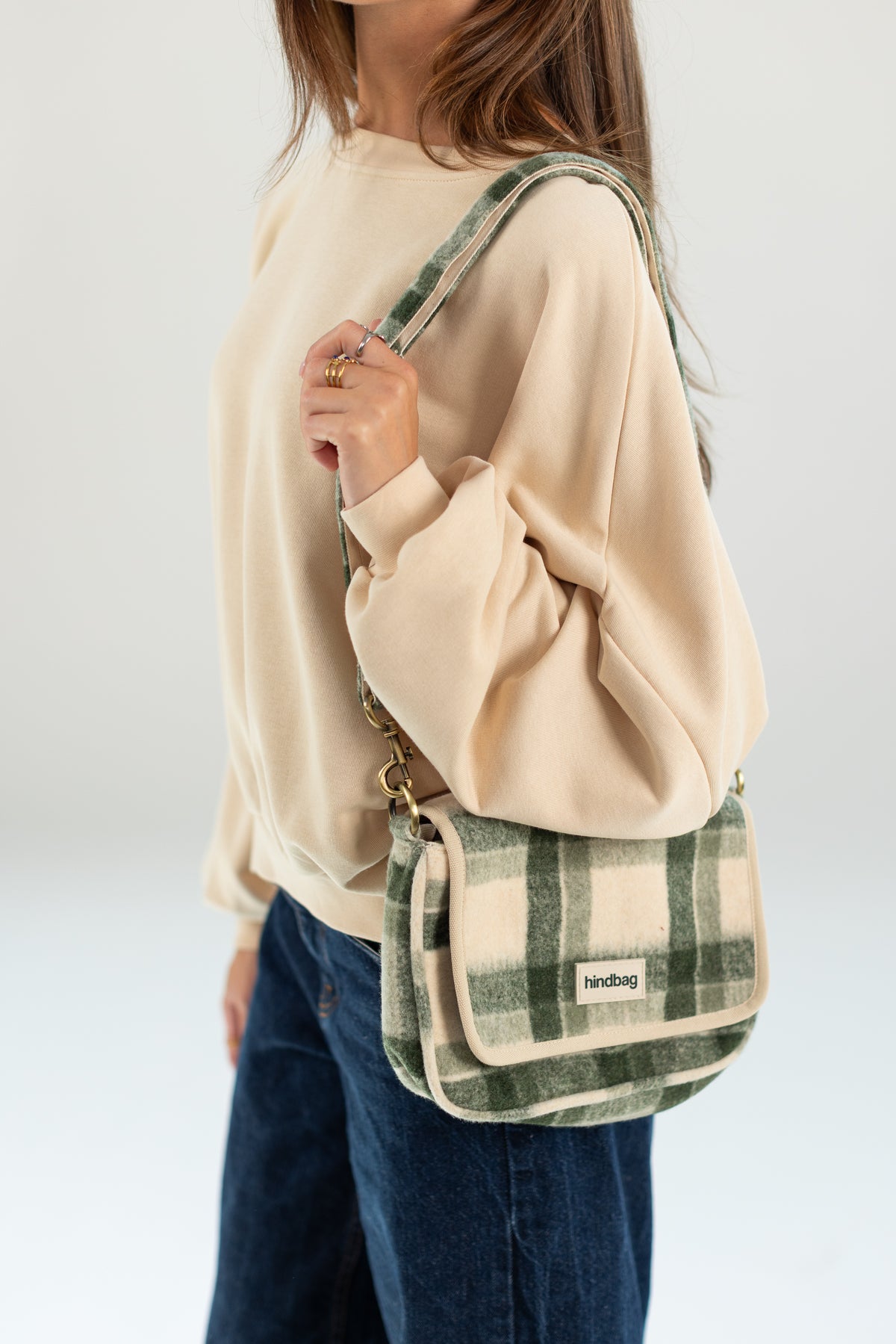 A woman wearing a cream sweatshirt and blue jeans carries a green and beige plaid crossbody bag with a small white brand label that reads 'klockdoo'.