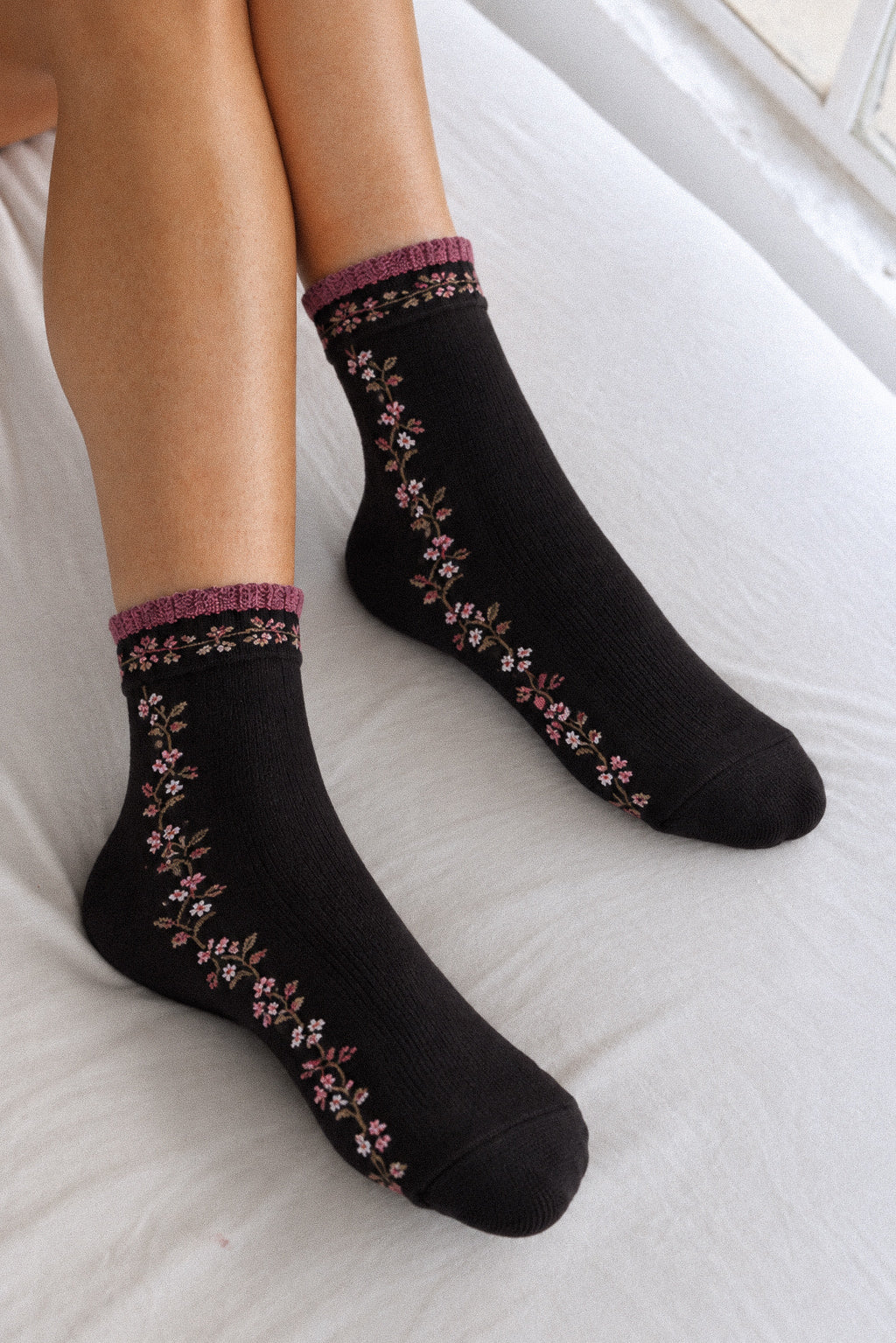 Black crew socks with pink and white floral embroidery and a purple cuff, worn on feet resting on white bedding.