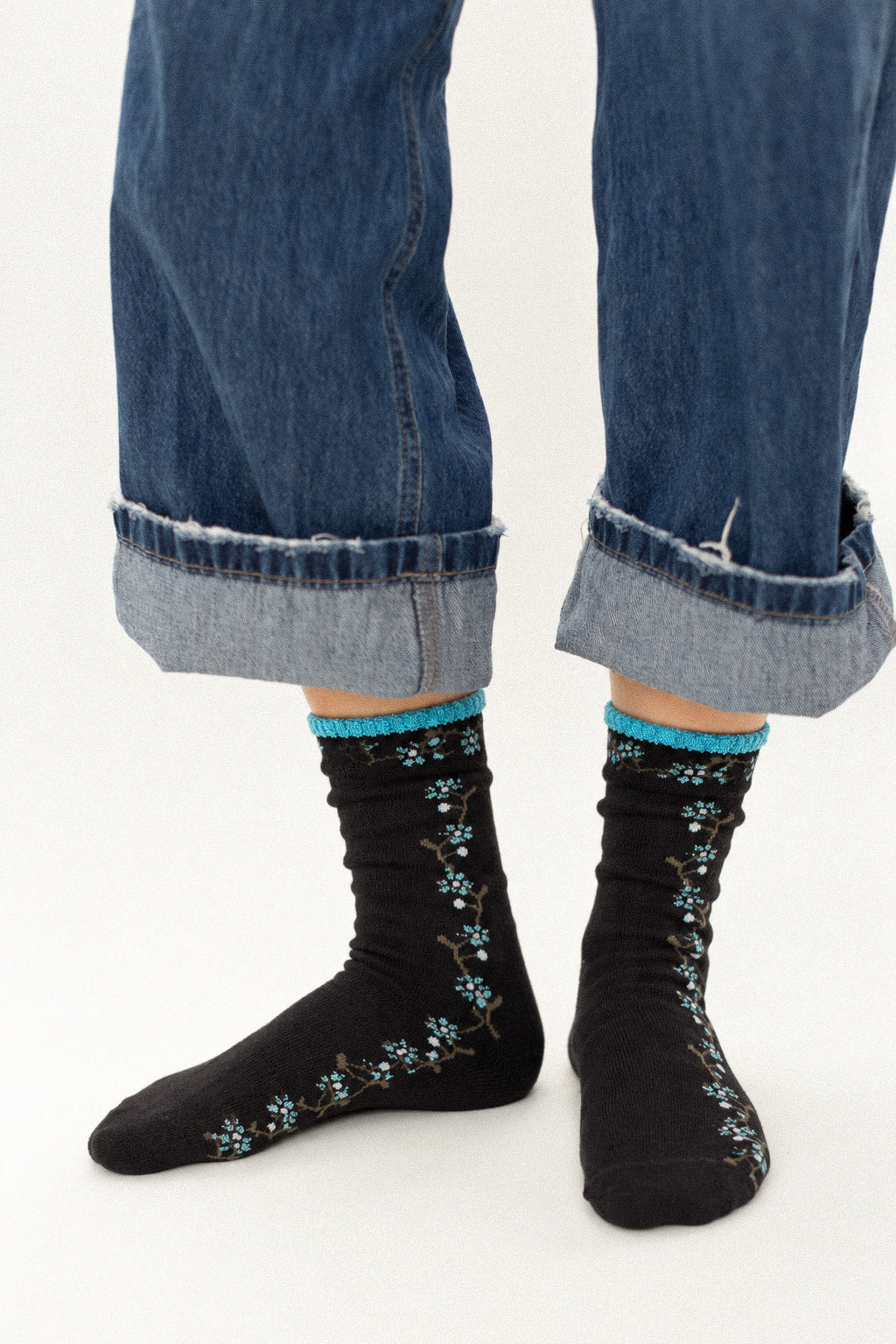 Black crew socks with a blue floral branch pattern and bright blue trim, worn with cuffed blue jeans against a white studio background.