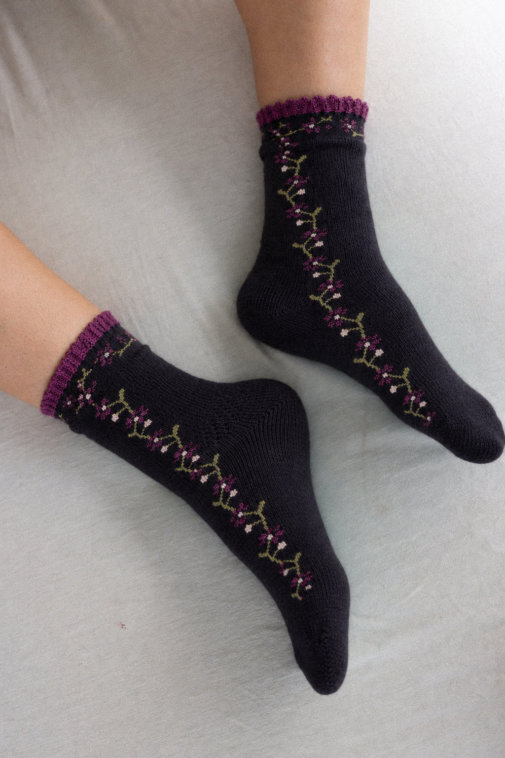 A pair of black crew socks with purple and green floral embroidery and purple ruffled cuffs, shown worn on feet against a light gray fabric background.