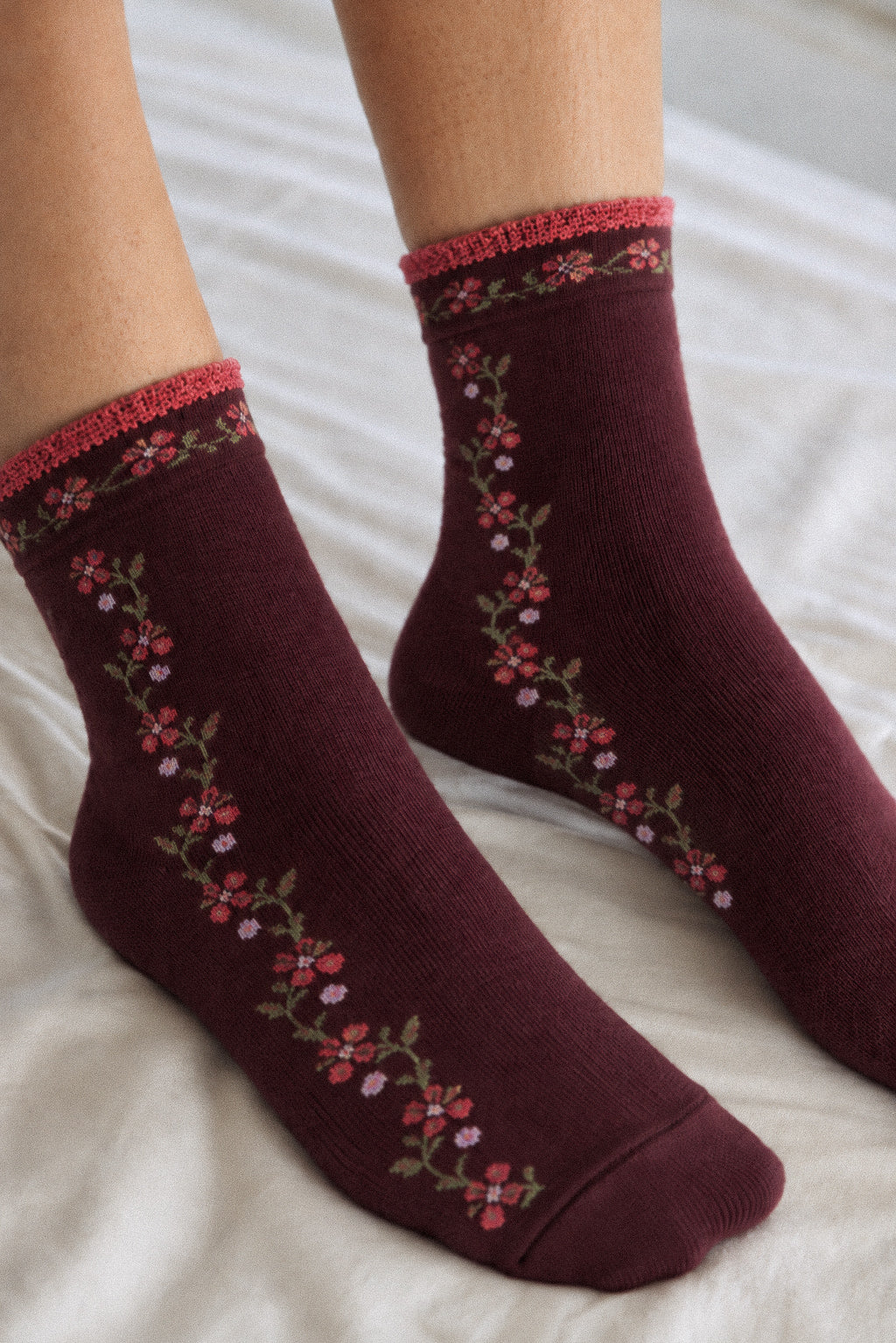 A pair of crew socks worn on feet, featuring a deep burgundy base with a vertical floral vine pattern in pink, red, and green, and a matching floral patterned ribbed cuff.