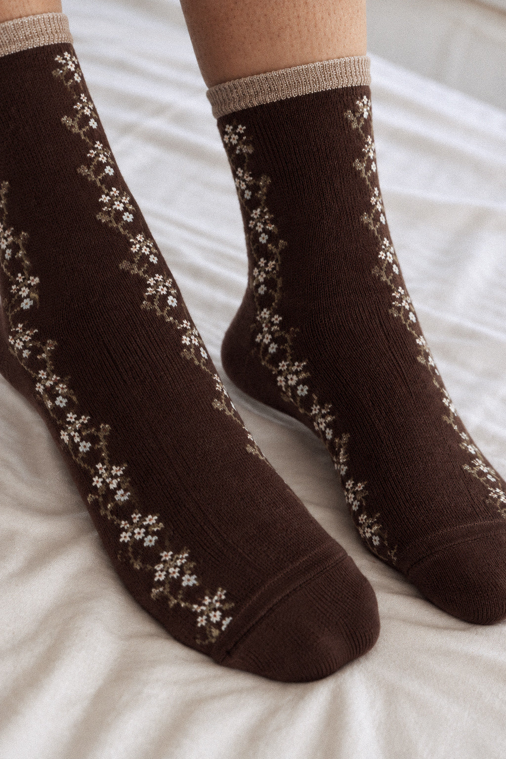 A close-up of a person's feet wearing dark brown crew socks with a beige and white floral pattern, resting on white bedding.