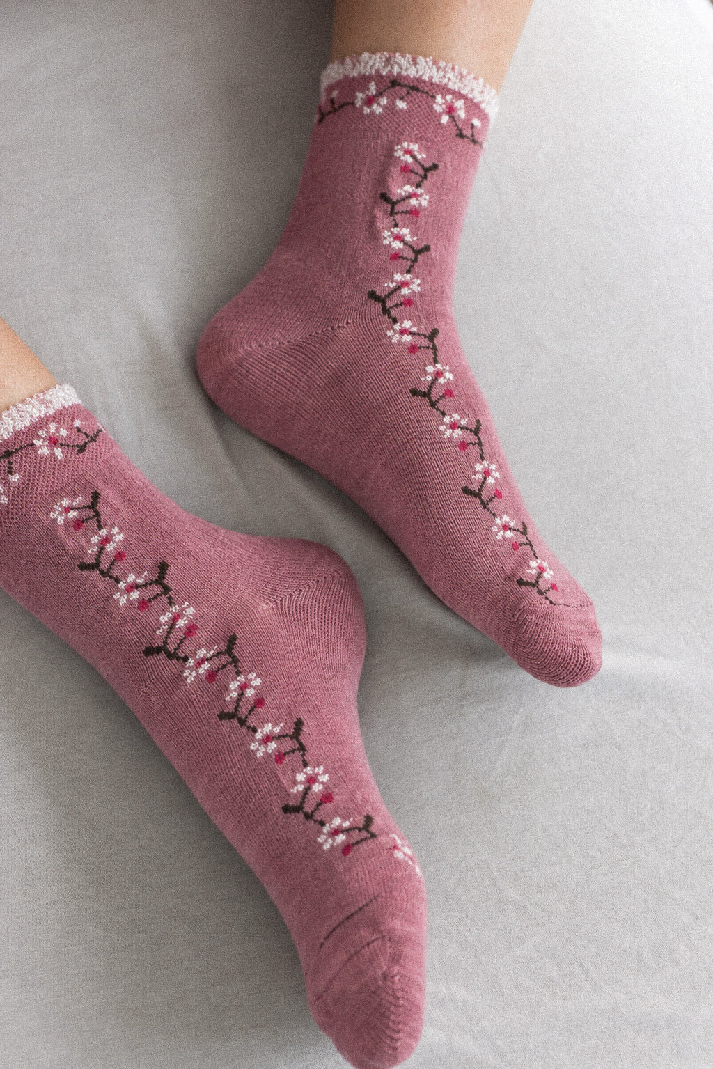A pair of dusty rose crew socks with a floral branch pattern in pink, white, and black, worn on feet resting on a light gray fabric surface.