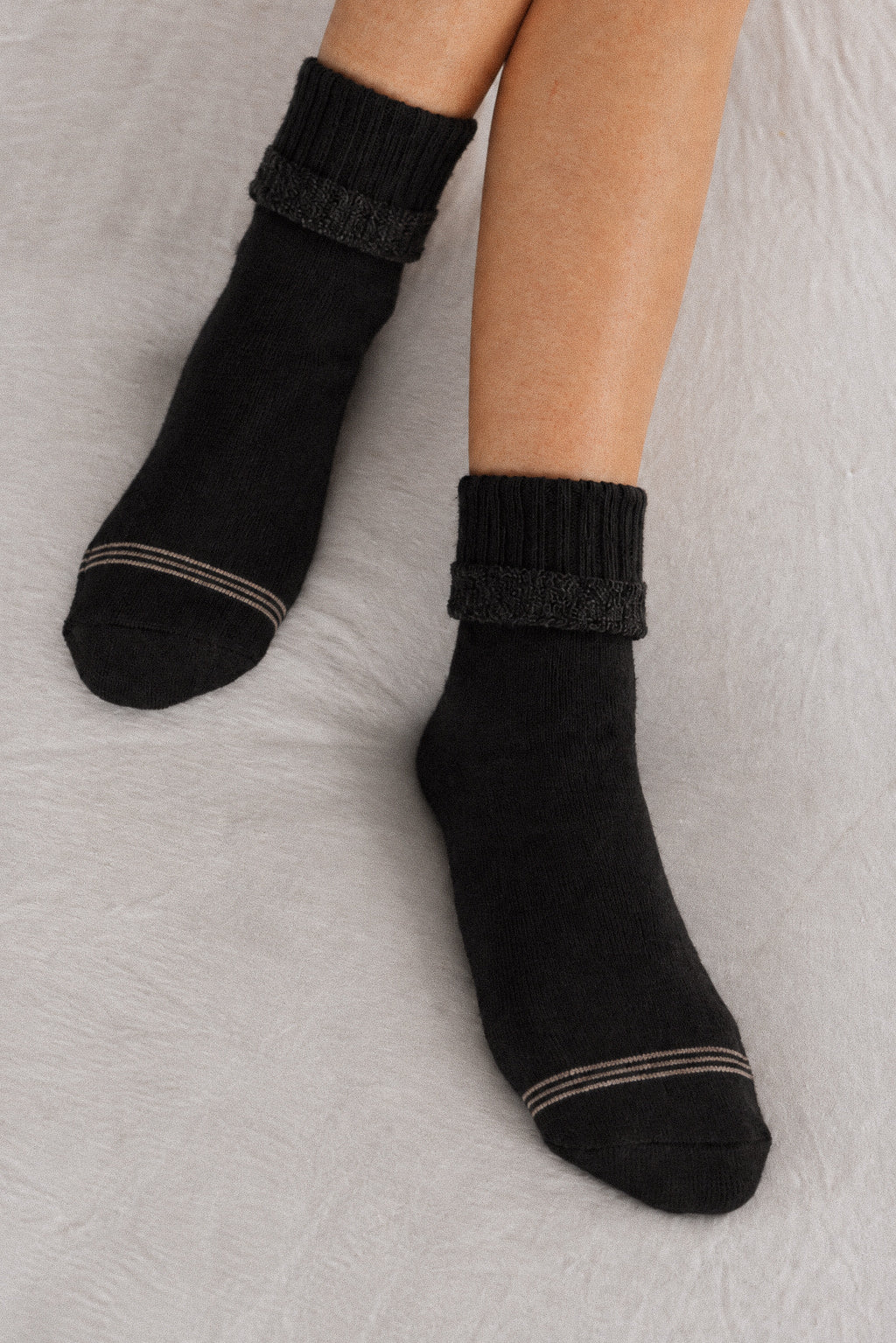 A pair of black ankle socks with ribbed cuffs and thin beige stripes near the toes, shown on a person's feet against a light gray textured background.
