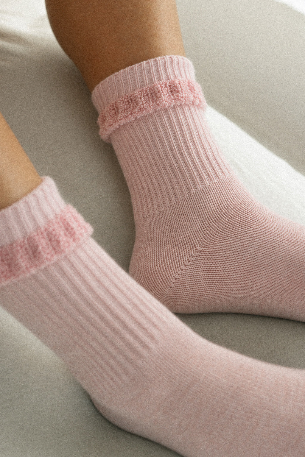 A close-up of a person's feet wearing light pink ribbed ankle socks with a textured lace-like band at the top, resting on a neutral gray surface.