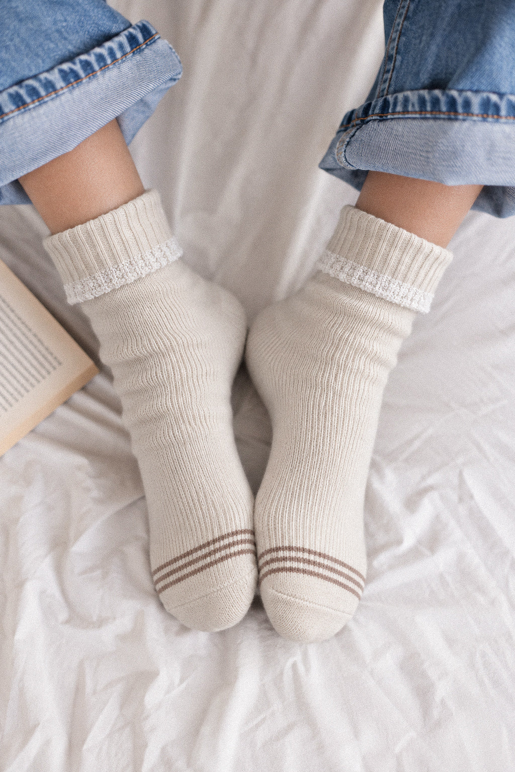 A person wearing cream-colored ribbed ankle socks with brown stripes near the toes, paired with cuffed blue jeans, sitting on a white bed.