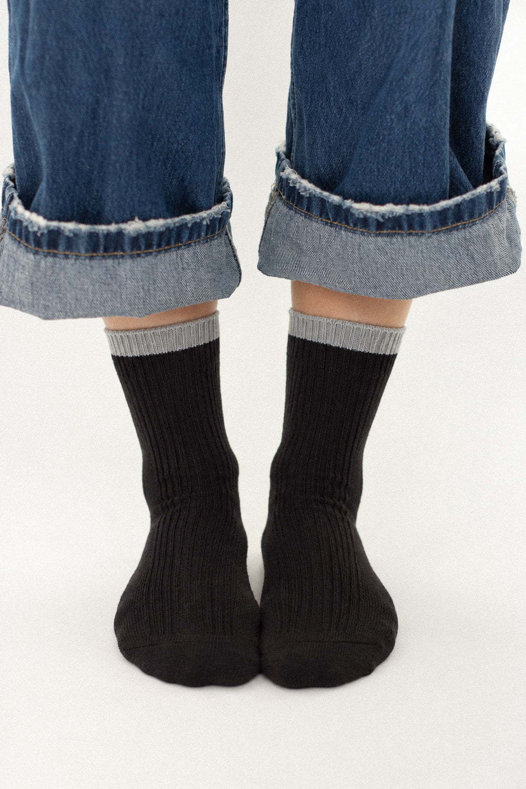 A close-up of a person's feet wearing black ribbed crew socks with a light gray ribbed cuff, paired with cuffed blue jeans on a white studio background.