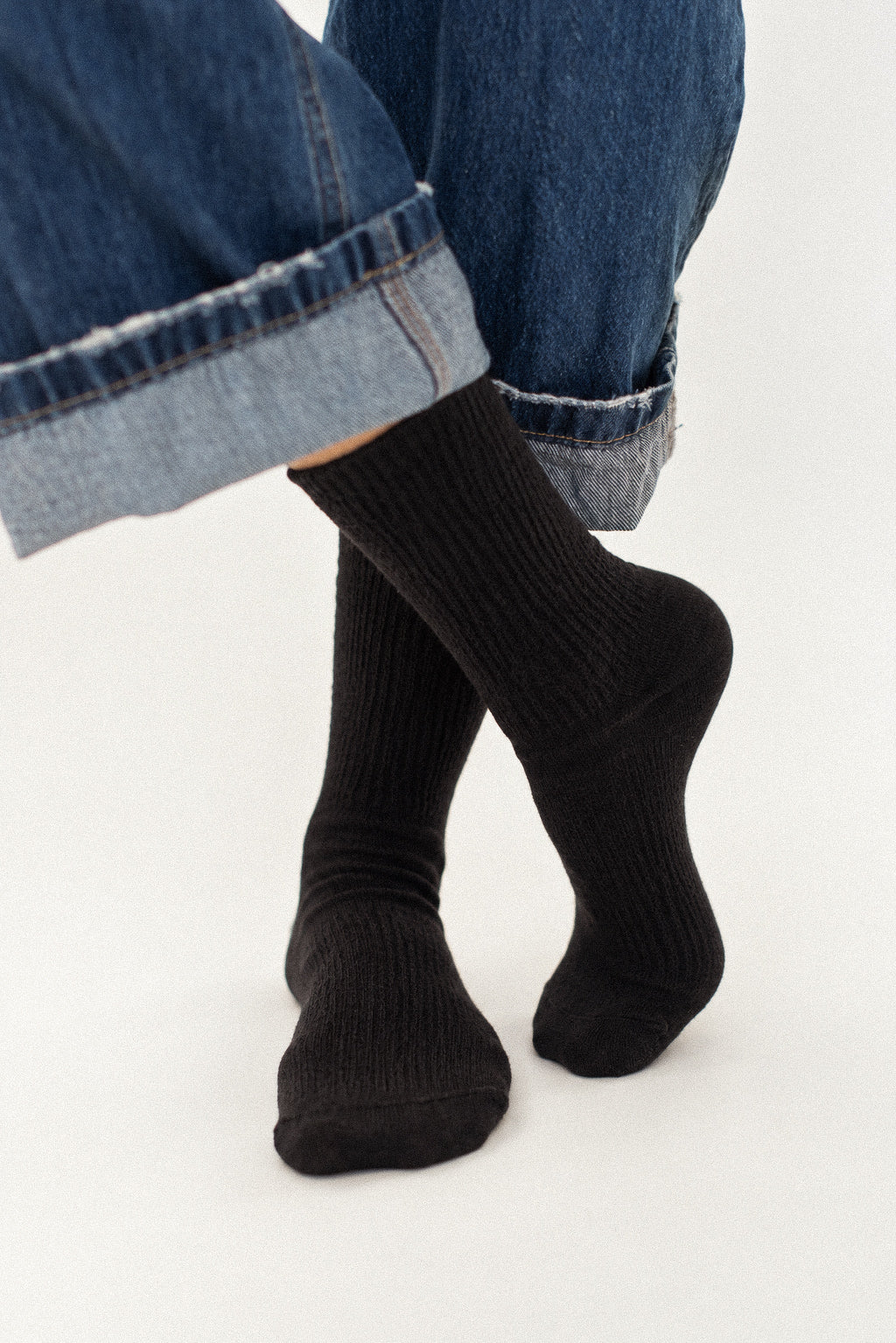 A close-up of a person's feet wearing black ribbed crew socks with cuffed blue jeans against a plain white background.