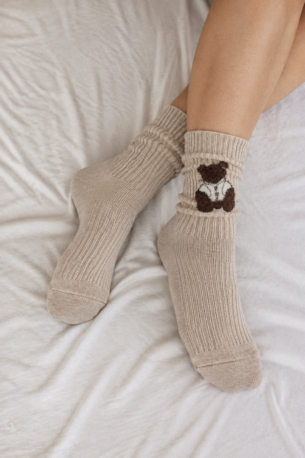A person's legs resting on white bedding, wearing ribbed beige crew socks with a small brown teddy bear graphic on the cuff.