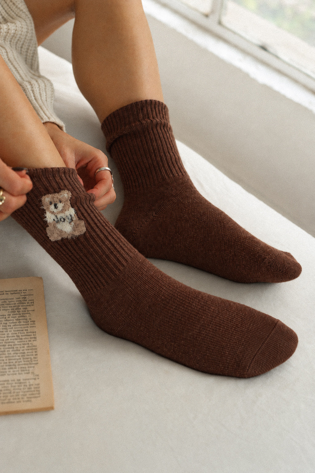 A person wearing brown ribbed crew socks, with one hand adjusting the cuff of one sock. The sock features a small embroidered teddy bear patch with the word 'love' written on it.
