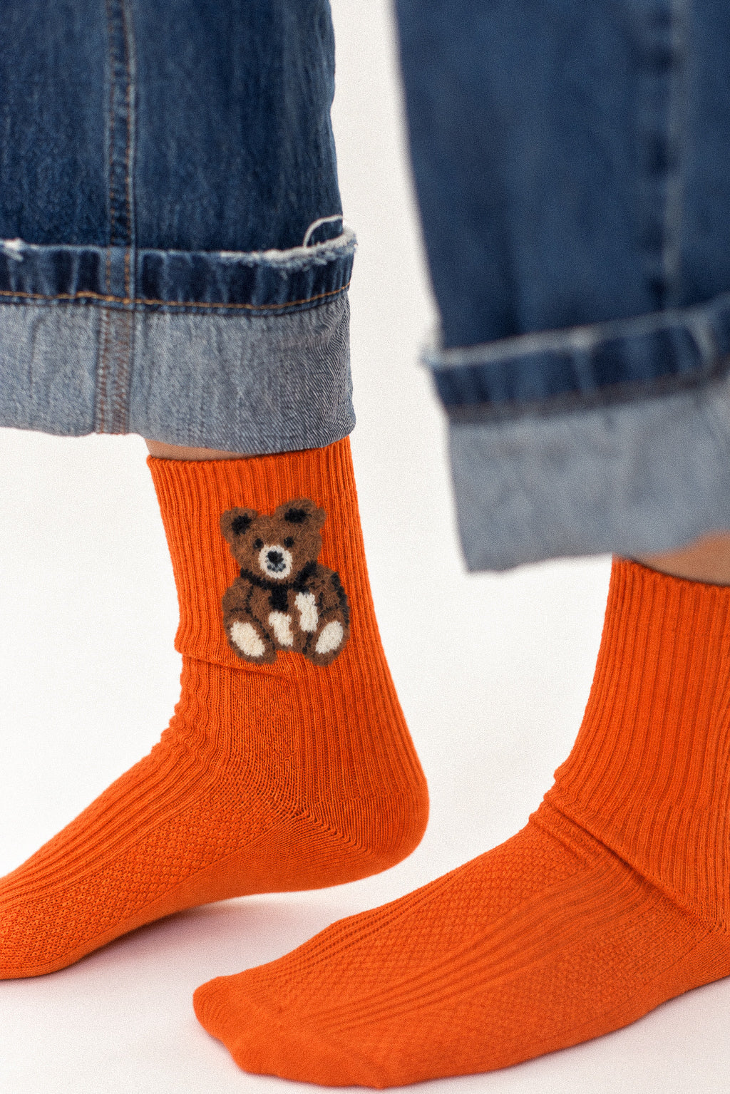 Bright orange ribbed crew socks worn with cuffed blue jeans. One sock features a small embroidered brown teddy bear with white details.