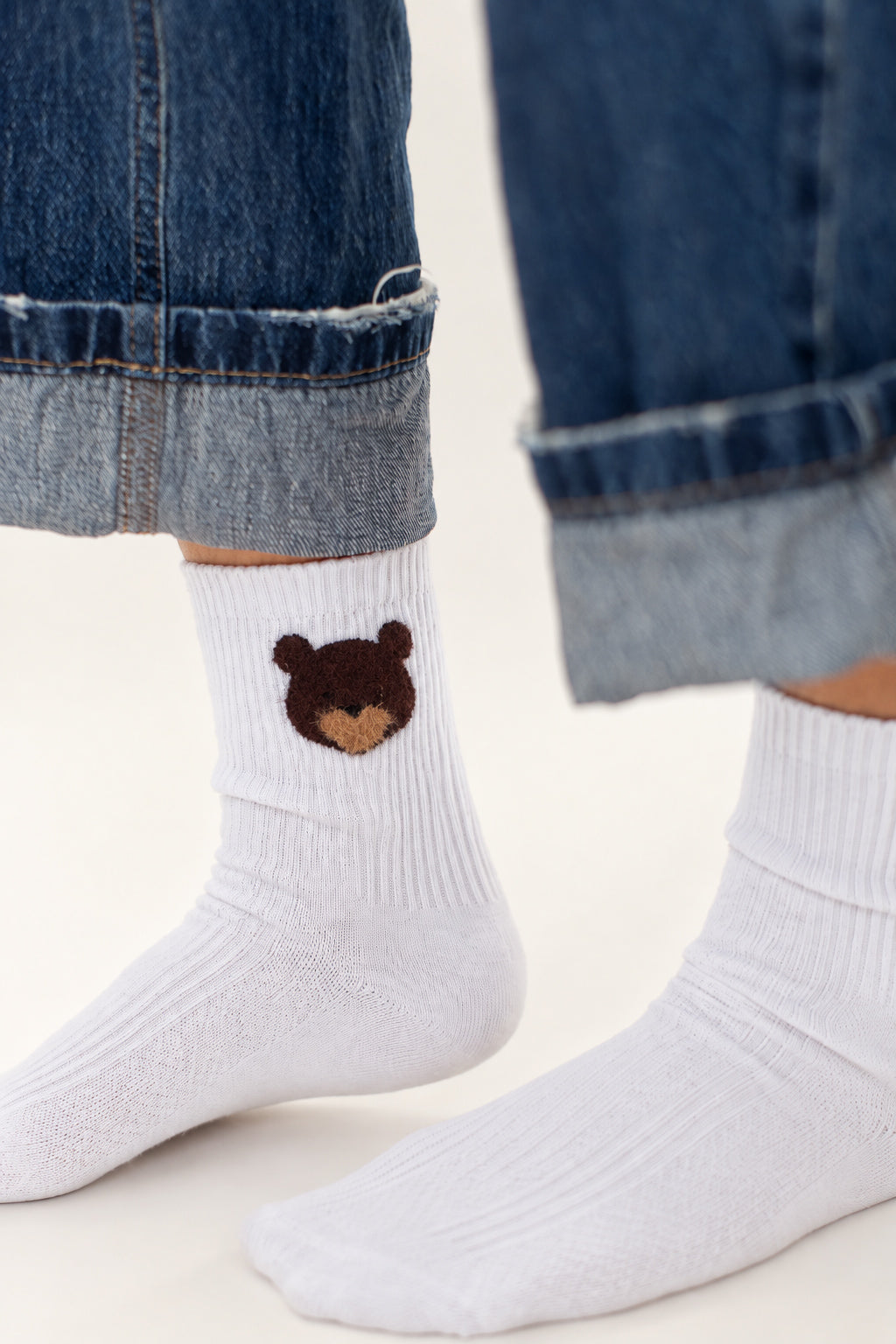 White ribbed ankle socks worn with cuffed blue jeans. One sock features a small embroidered brown bear face with a tan snout.