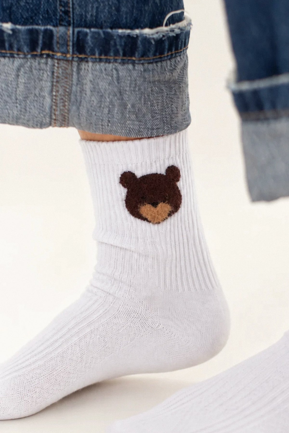White ribbed crew socks worn with cuffed blue jeans, featuring a small brown bear face motif on the side.