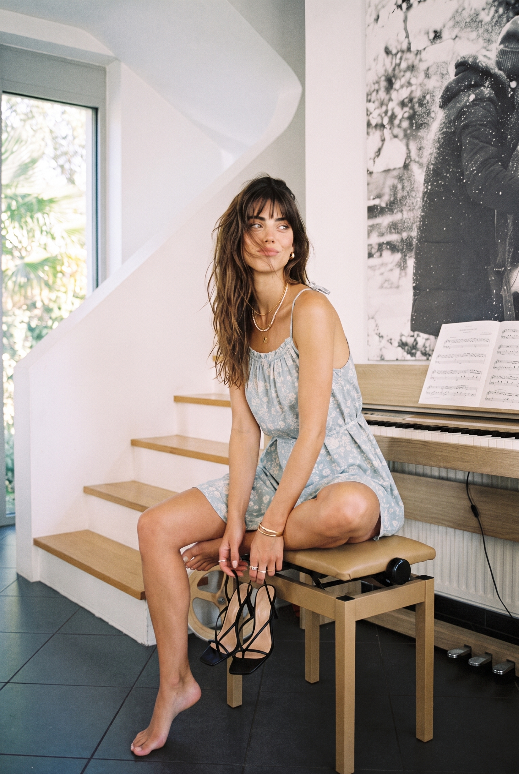A woman sits on a wooden bench in a bright, modern room with a spiral staircase and a piano. She is wearing a light blue and white patterned sleeveless dress and black strappy high heels.