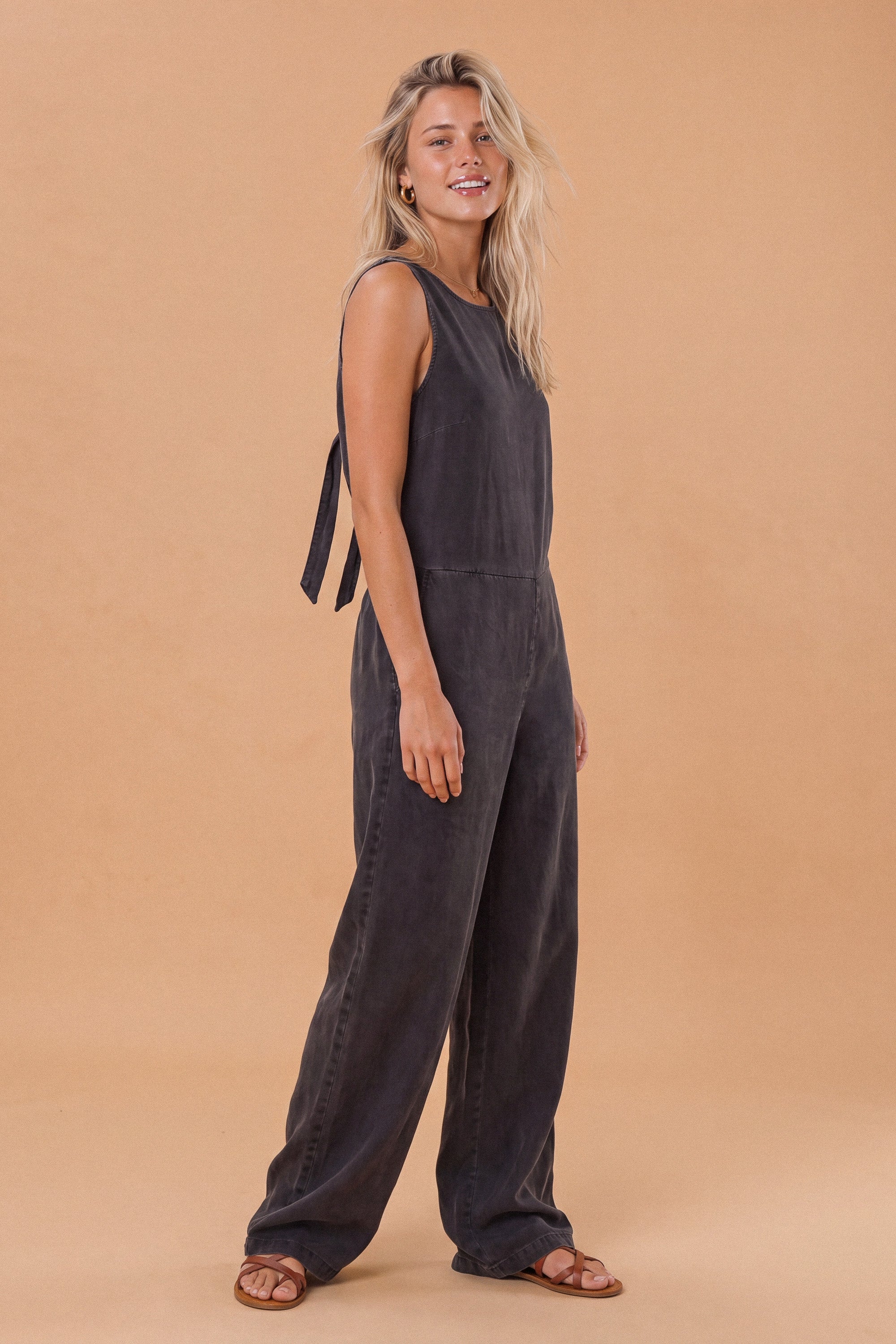 A woman modeling a dark charcoal gray sleeveless wide-leg jumpsuit with tie details on the shoulders, standing against a solid warm beige studio background.