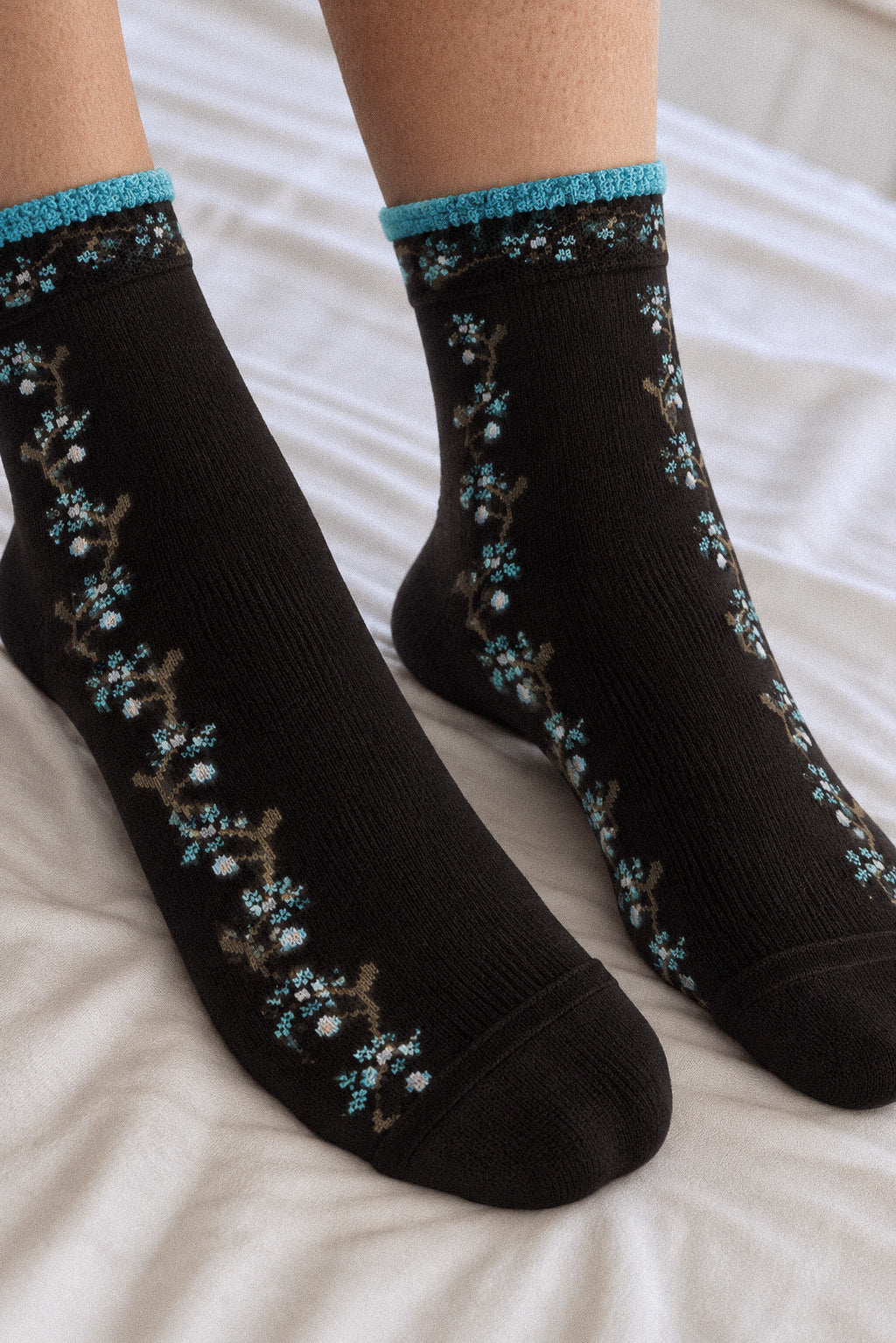 A pair of black crew socks with a floral branch pattern in light blue and brown, featuring a bright blue ribbed cuff, shown on a person's feet resting on white bedding.