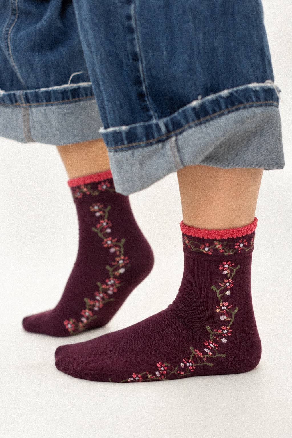 A person wearing dark burgundy crew socks with a pink floral pattern and a pink ribbed cuff, paired with cuffed blue jeans.