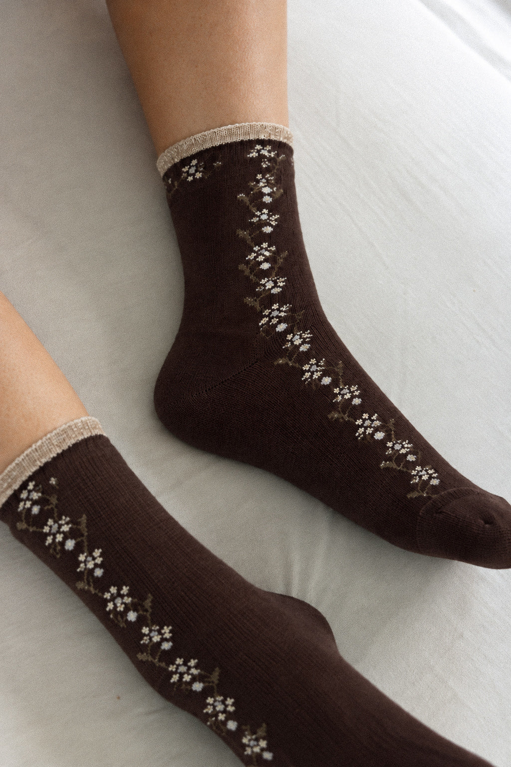 A pair of dark brown ankle socks with a cream and white floral knit pattern, worn on feet resting on a light-colored fabric surface.