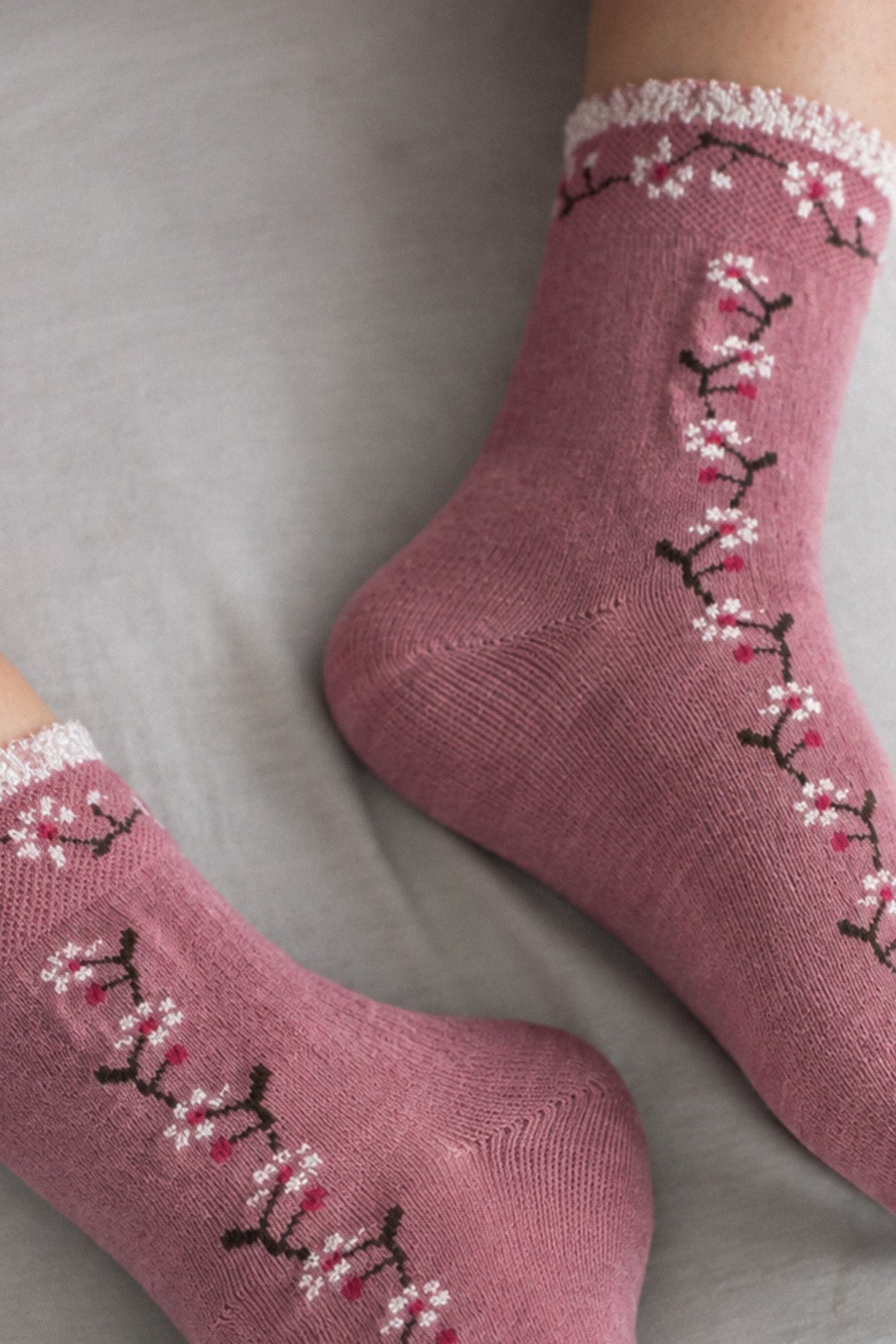 A pair of dusty rose knit socks with a white floral branch pattern and white ruffled trim, shown worn on feet against a neutral gray background.