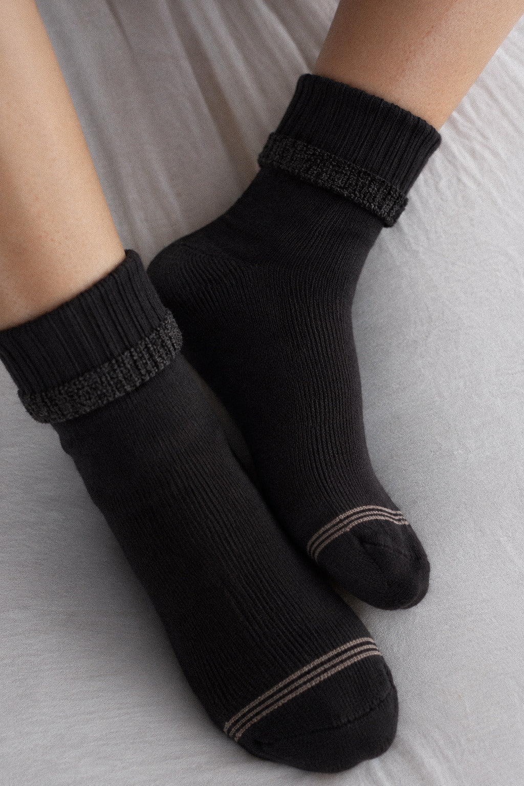 A pair of black crew socks with textured gray ribbed cuffs and thin beige stripes near the toes, shown worn on feet against a light gray fabric background.