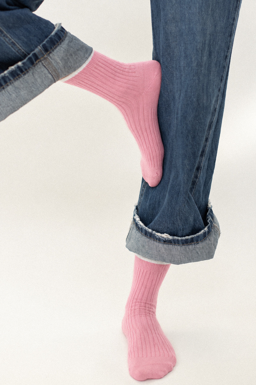 A close-up of a person's legs wearing pink ribbed crew socks and cuffed blue jeans against a plain light background.