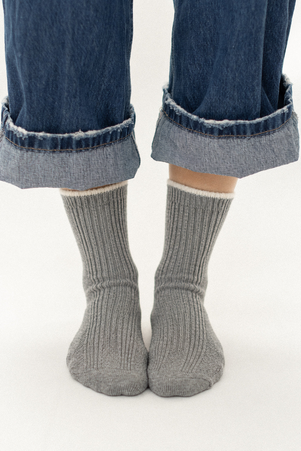 A close-up of a person's feet wearing gray ribbed crew socks with a white cuff, paired with cuffed blue jeans, against a plain white background.