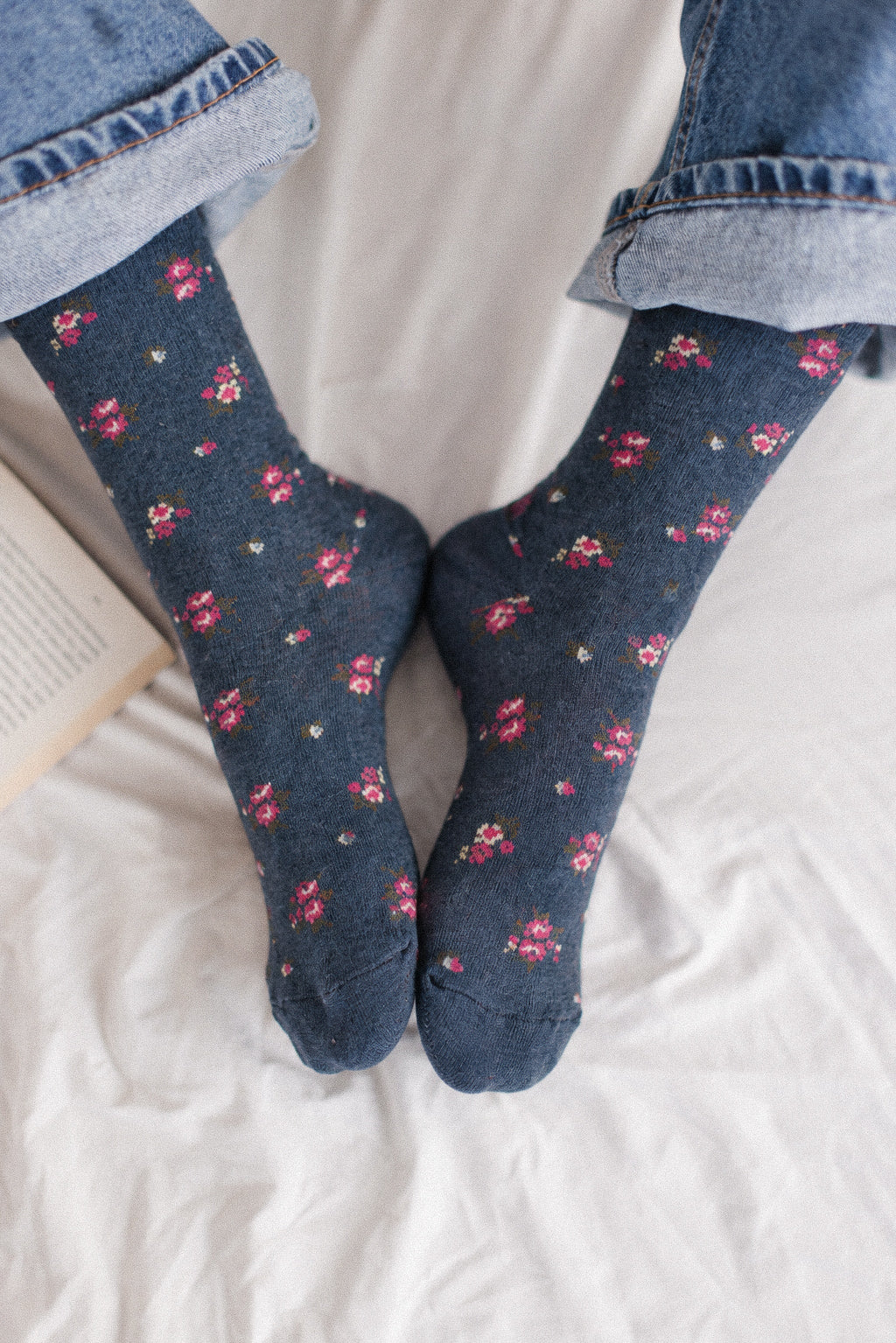A pair of dark blue socks with a small pink and red floral pattern, worn with cuffed blue jeans on white fabric.