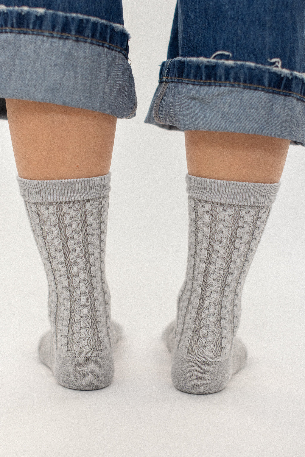 A pair of light gray cable-knit crew socks worn on feet, shown from the back with cuffed blue denim jeans against a plain white background.