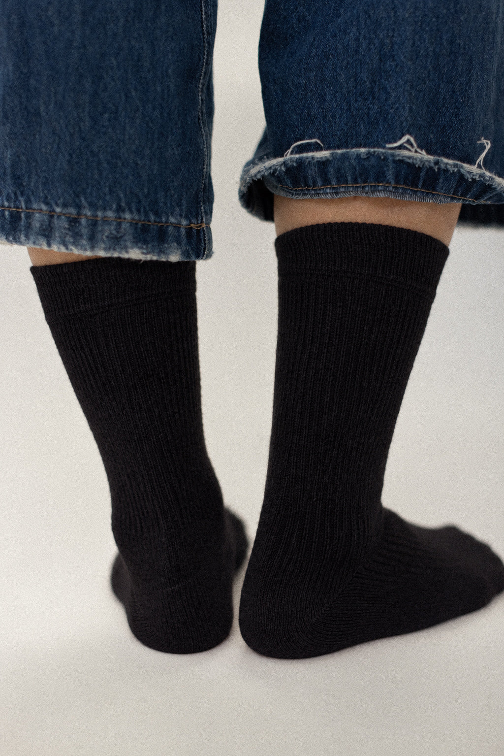 A close-up shot of a person's feet wearing black ribbed crew socks with blue denim jeans, shown from the back against a light gray background.