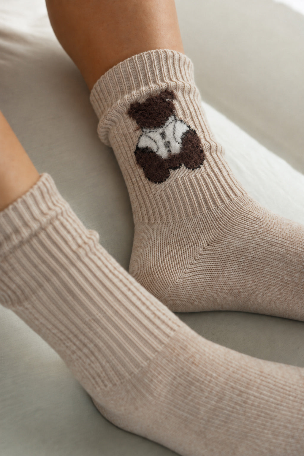Close-up of a person wearing ribbed ankle socks in a light beige color, with one sock featuring a knitted brown teddy bear holding a white heart.