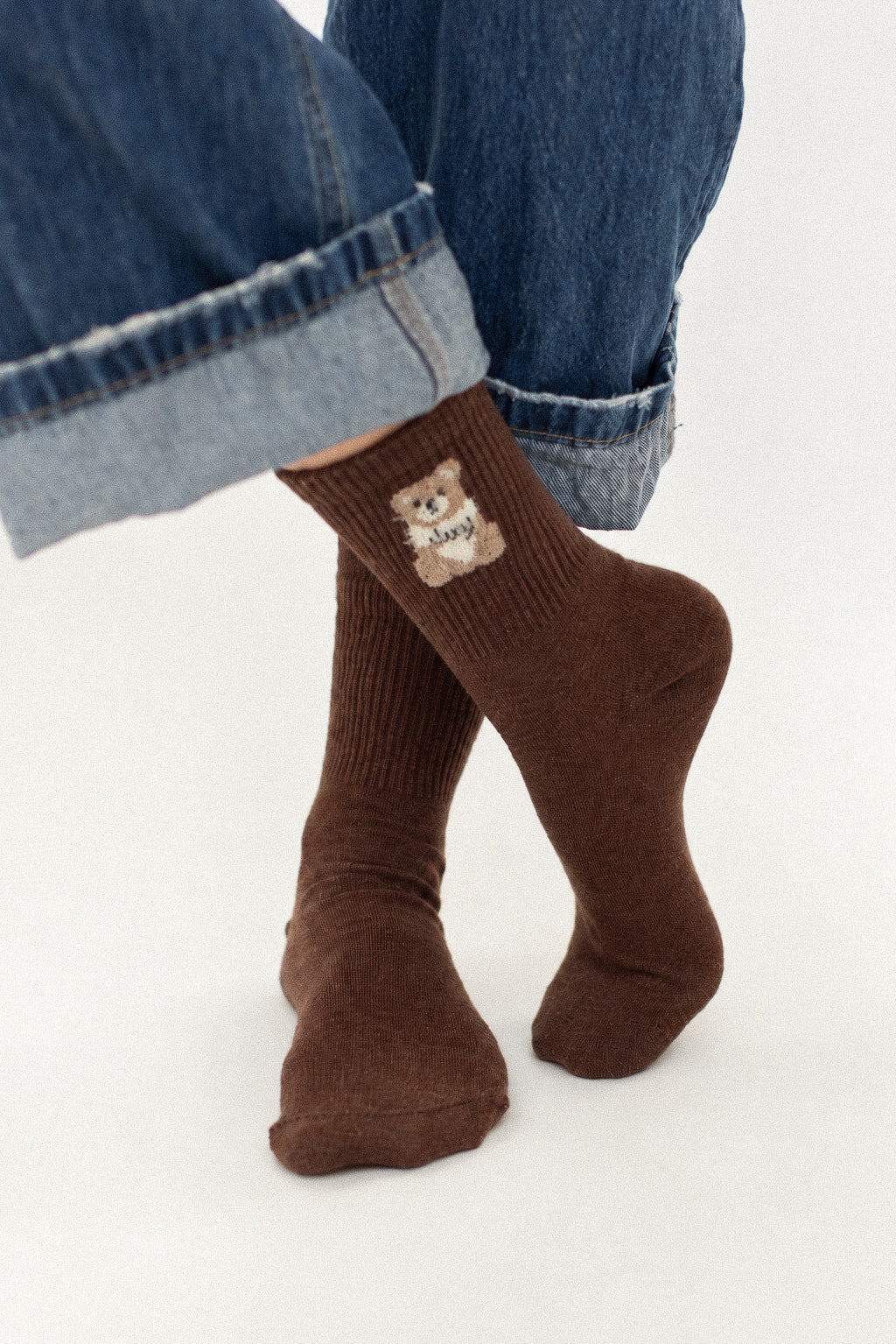 A person wearing brown ribbed crew socks with a small teddy bear graphic on the side, paired with cuffed blue jeans against a plain white background.