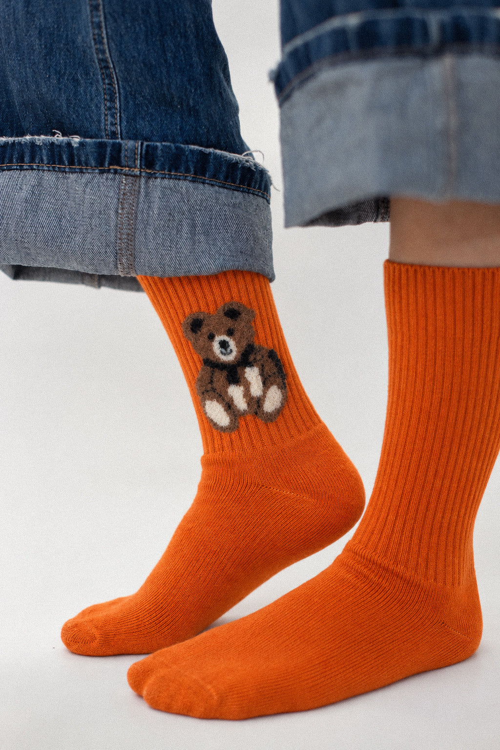 Bright orange ribbed crew socks worn with cuffed blue denim jeans. One sock features a small embroidered brown teddy bear with white details.